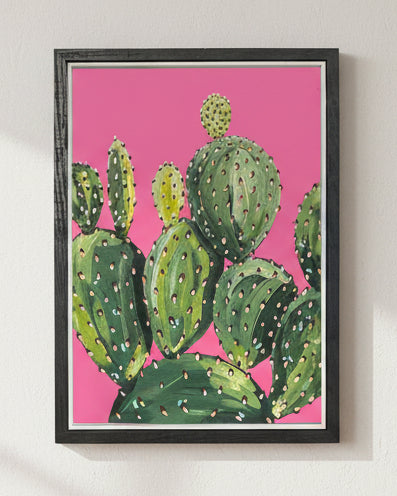 A framed art print featuring a painted green prickly pear cactus on a bright pink background, set in a black frame.