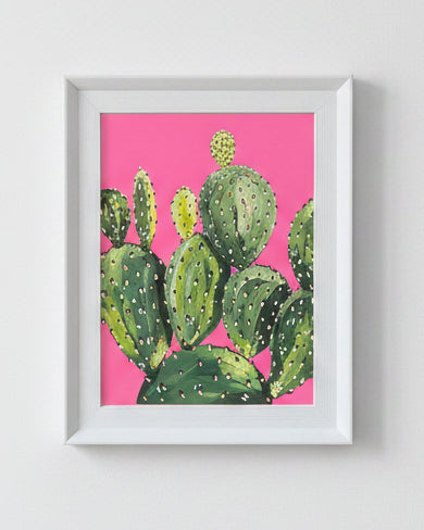A framed art print featuring a green prickly pear cactus painted against a vibrant pink background, housed in a white frame.